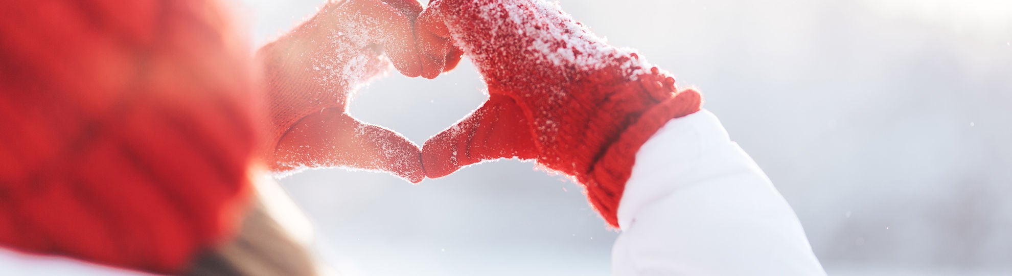 Zwei Hände in roten Winterhandschuhen formen ein Herz im Schnee – Symbol für Herzenswärme und Schutz in der kalten Jahreszeit, Bildquelle Anastassiya