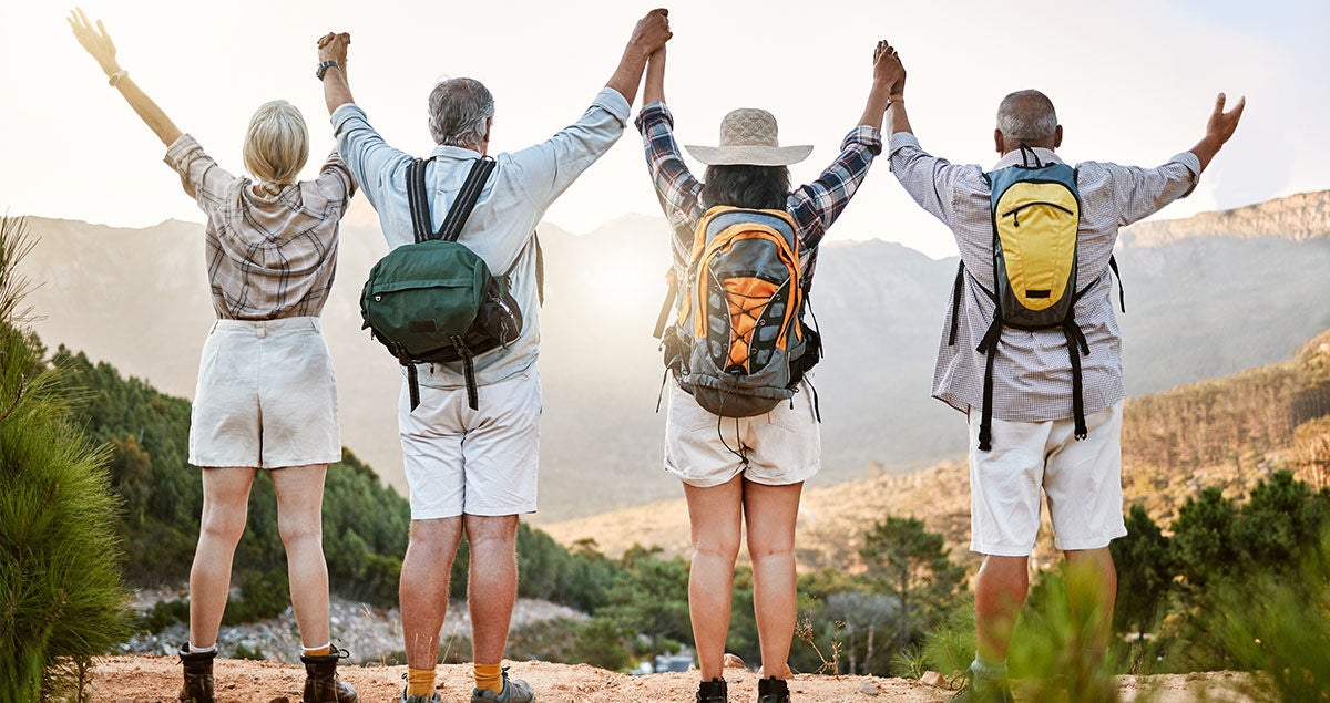 Vier Seniorinnen und Senioren, die Wanderoutfits tragen, stehen nebeneinander auf einem Berg, fassen sich an den Händen, strecken die Arme nach oben und genießen die Aussicht. Bildquelle: Adobe Stock / Courtney Haas/peopleimages.com