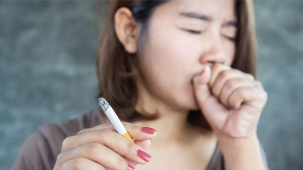 Eine junge Frau hält eine brennende Zigarette in der Hand und  hustet. iStock/Doucefleur 