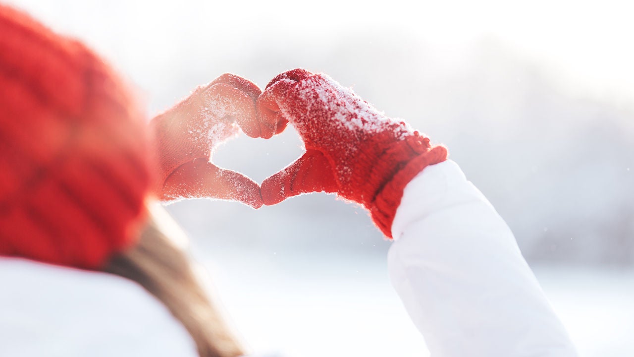 Zwei Hände in roten Winterhandschuhen formen ein Herz im Schnee – Symbol für Herzenswärme und Schutz in der kalten Jahreszeit, Bildquelle Anastassiya