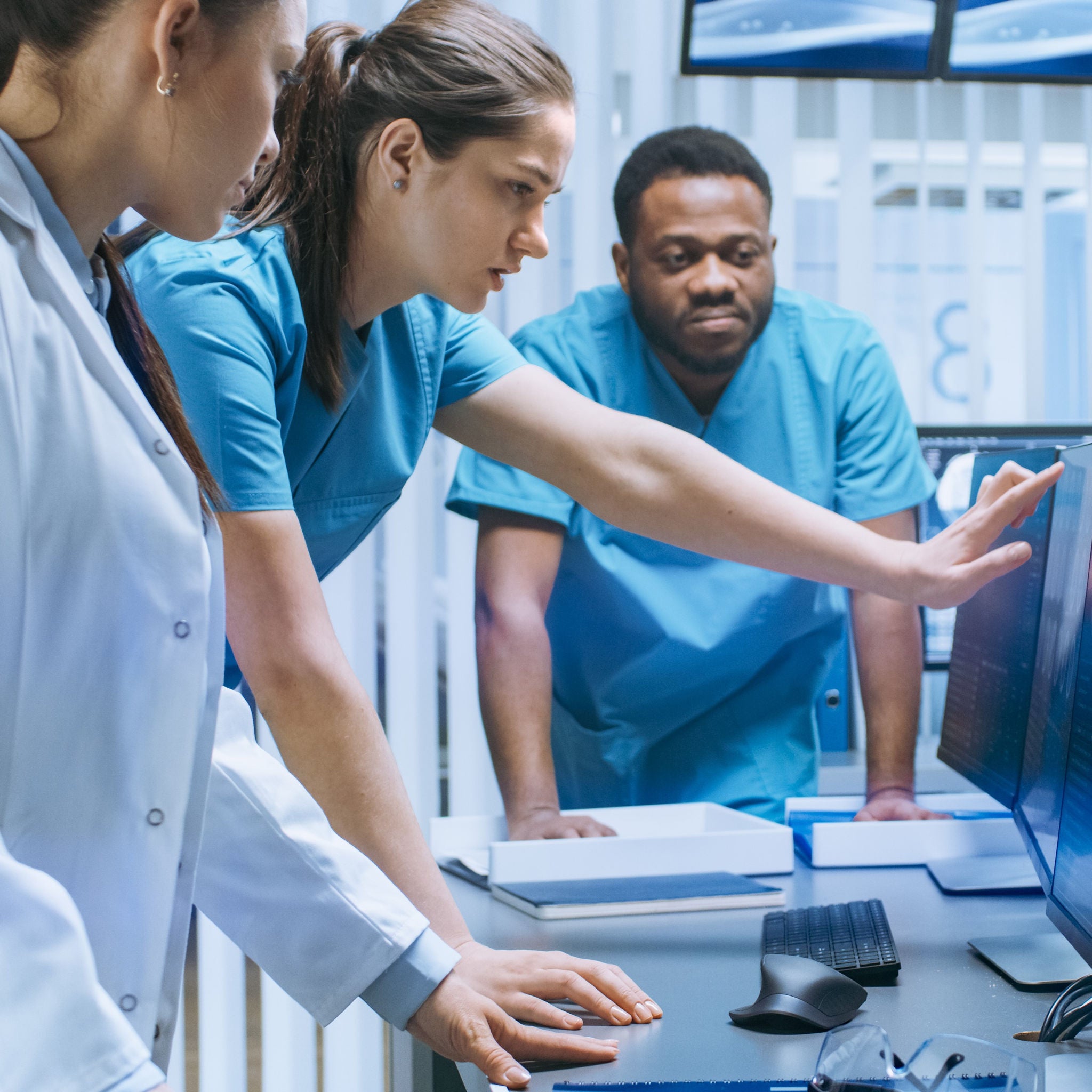 Drei junge Mediziner in Klinik-Kleidung stehen an einem Schreibtisch und deuten auf einen Bildschirm